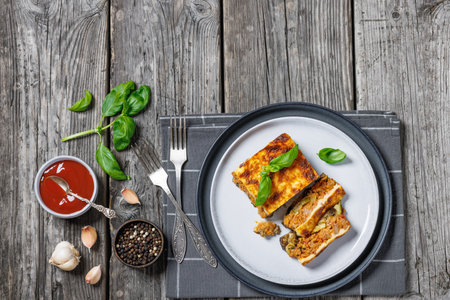 Moussaka, layer of rich tomato beef sauce on fried eggplant topped with bechamel sauce on plate on rustic wooden table with forks and basil, Greek recipe, horizontal view from above, flat layの写真素材