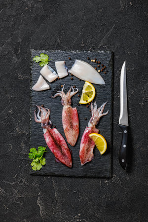 fresh raw baby squid with lemon and parsley on black slate plate with knife on concrete table, horizontal view from above, flat layの写真素材