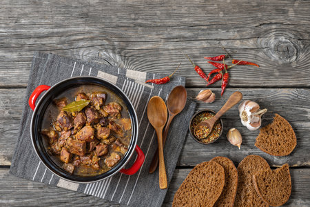 beer braised pork stew in red pot on rustic wooden table with spoons, seasoning, rye bread, dry red peppers, garlic, horizontal view from above, flat lay, free spaceの写真素材