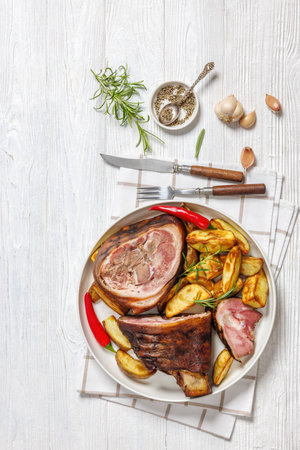 Smoked pork knuckle, ham hock with roasted potatoes, red chili peppers on whie platter on white wooden table with cutlery, horizontal view, flat lay, free spaceの写真素材