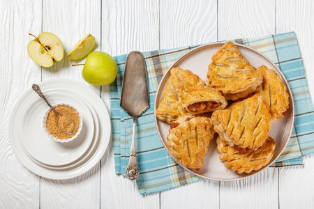puff pastry apple turnovers on a plate on white wooden table with plates, napkin, cake shovel and apples, horizontal view from above, flat layの写真素材