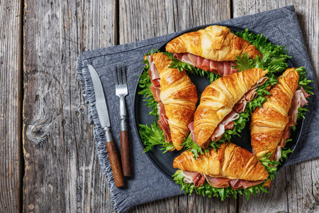 croissant sandwiches with ham and fresh lettuce on a plate on rustic wooden table with napkin and cutlery, horizontal view from above, flat lay, free spaceの写真素材