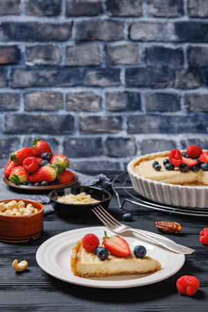 Piece of Berry tart with marzipan based crust and coconut almond cream filling on plate on wooden table with ingredients, fresh berries, brick wall in background, vertical view, close-upの写真素材