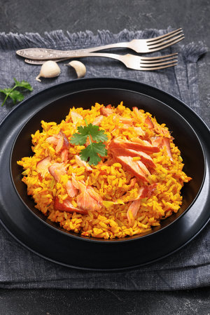 hot smoked salmon kedgeree, spiced cooked rice with salmon file flakes in black bowl on black concrete table with forks, garlic, vertical view from aboveの写真素材