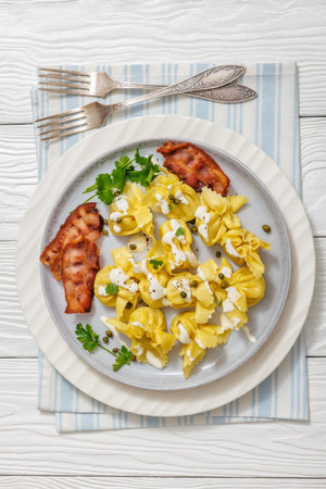 sacchettoni, italian pasta with bacon and cheese filling drizzled with white sauce and sprinkled with caper on plate on white wooden table with forks, horizontal view, flat lay, close-upの写真素材