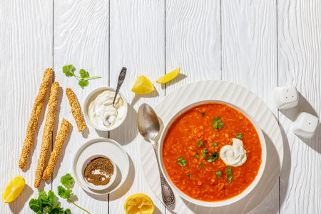 Red Lentil Tomato Soup with sour cream and cilantro in white bowl on white wooden table with spoon, lemon, grissini and ground cumin, horizontal view from above, flat lay, free spaceの写真素材