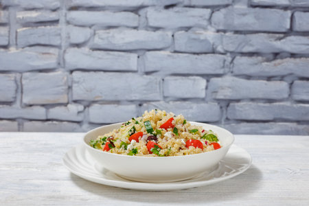Quinoa salad with tomatoes, kalamata olives, cucumber, crumbled goat cheese and red onion in a white bowl on white wooden table in front of grey brick wall, horizontal view from aboveの写真素材