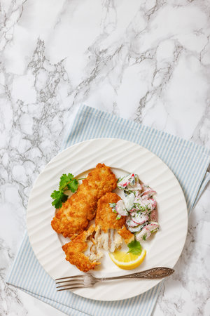 fried battered white fish fillets with creamy radish cucumber salad on white plate with fork on marble table, vertical view from above, free spaceの写真素材
