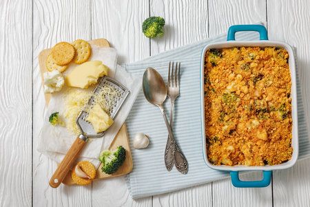 broccoli cauliflower cheese casserole with crumble topping in a baking dish on a white wooden table with grated cheese, garlic, and crackers, horizontal view from above, flat layの写真素材
