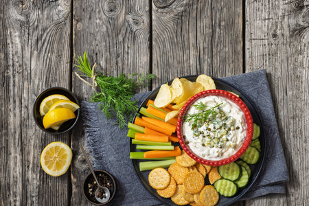 creamy yogurt pickles caper dill dip in red bowl on platter with carrot and celery sticks, cucumber, potato chips and crackers on grey wood table, horizontal view from above, flat lay, free spaceの写真素材