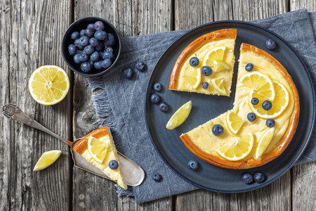Cheesecake with blueberries and lemon sliced on a plate with cake shovel and fresh berries on rustic wooden table, horizontal view from above, flat layの写真素材