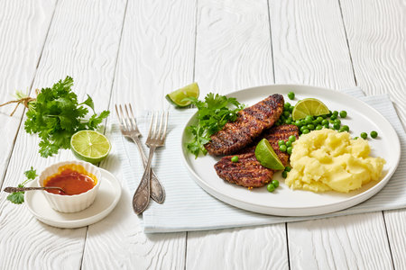spicy grilled beef cutlets with potato mash and green peas on a plate on white wooden table with coriander, lime and hot sauce, horizontal view from aboveの写真素材