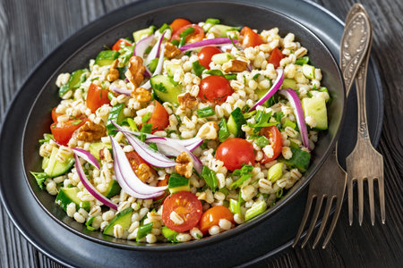 Barley salad with cucumber, tomatoes, red onion and walnuts in a black bowl with forks on black wooden table, close-up, dutch angle viewの写真素材
