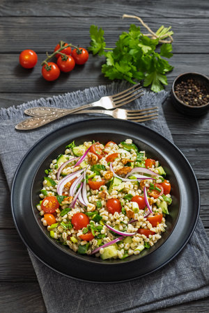 Barley salad with cucumber, tomatoes, red onion and walnuts in a black bowl on dark wooden table with forks and parsley, vertical view from aboveの写真素材