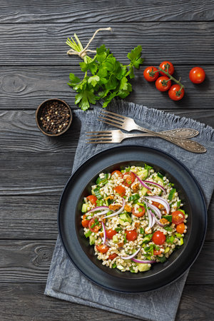 Barley salad with cucumber, tomatoes, red onion and walnuts in a black bowl on dark wooden table with forks and parsley, horizontal view from above, flat lay, free space on topの写真素材