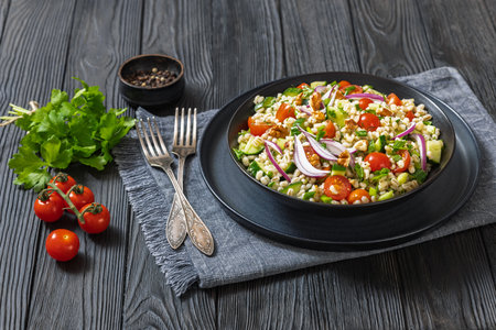 Barley salad with cucumber, tomatoes, red onion and walnuts in a black bowl on dark wooden table with forks and parsley, vertical view from aboveの写真素材