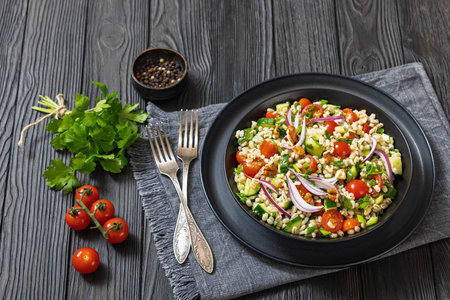 Barley salad with cucumber, tomatoes, red onion and walnuts in a black bowl on dark wooden table with forks and parsleの写真素材