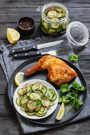 golden brown roast chicken leg with crispy skin, served on a plate on black wooden table with marinated cucumber salad, vertical view from above, close-upの写真素材