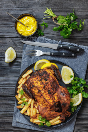 whole crispy skin roasted chicken on a platter with fried potatoes, lemon wedges and cilantro on dark wooden table with mustard, napkin and cutlery, vertical view from aboveの写真素材
