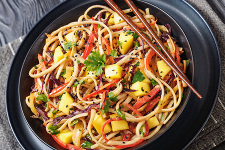 Thai Mango Noodle Salad with pepper, red cabbage, mint and cilantro, peanut sauce dressing in a black bowl with chopsticks on black wooden table, dutch angle viewの写真素材
