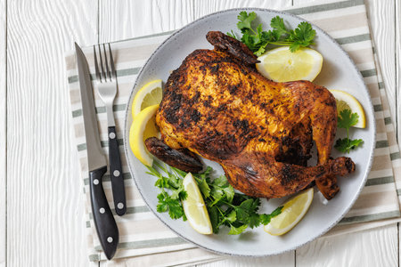whole crispy skin roasted chicken on a platter with lemon wedges and cilantro on white wooden table with cutlery, horizontal view from above, flat lay, free spaceの写真素材