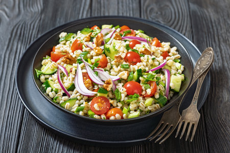 Barley salad with cucumber, tomatoes, red onion and walnuts in a black bowl with forks on black wooden table, close-upの写真素材