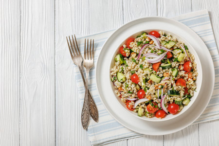 Pearl Barley salad with cucumber, tomatoes, red onion and walnuts in a white bowl with forks on white wooden table, horizontal view from above, flat lay, free spaceの写真素材