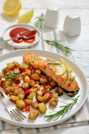 Vertical close-up of oven-baked salmon fillet with golden fried potatoes, lemon, and rosemary on a white plate atop a rustic white wooden table. Real photo, not AI-generatedの写真素材