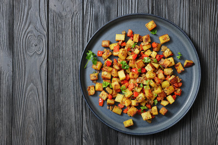 golden fried potatoes cubes with red bell pepper on a dark grey plate on a black wooden table, horizontal view from above, flat lay, free spaceの写真素材