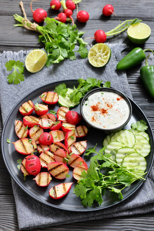 Grilled radishes with cucumber slices and sour cream sauce served on a dark grey plate on a black wooden table with fresh cilantro, jalapeno pepper, lime, vertical view, real photo, not AI generatedの写真素材