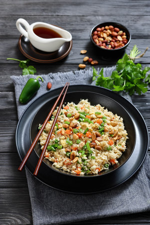 fried cauliflower rice with scrambled egg and veggies, topped with roasted peanut in in a black bowl on a black wooden table, with chopsticks, vertical view, not AI generatedの写真素材