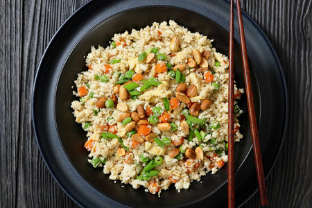 fried cauliflower rice with scrambled egg and veggies, topped with roasted peanut in in a black bowl on a black wooden table with chopsticks, flat lay, top view, close-up, not AI generatedの写真素材