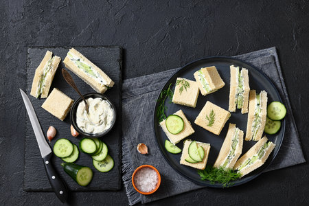 cucumber sandwiches layered with cream cheese spread on black plate with ingredients and knife on a black stone board, horizontal view from above, flat lay, real photo, not AI generatedの写真素材