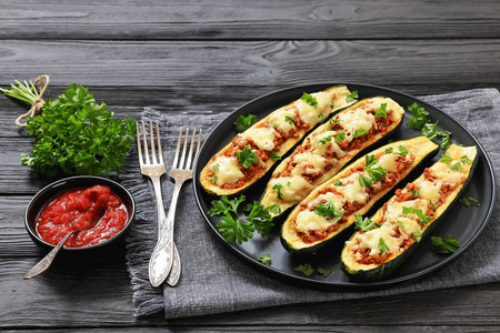 stuffed zucchini boats filled with hearty meat sauce topped with melted cheese and fresh parsley served on a plate on black wood table with tomato sauce and forks, horizontal view, not AI generatedの写真素材