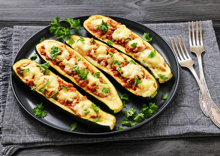 stuffed zucchini boats filled with bolognese sauce topped with melted cheese and fresh parsley served on a plate on black wood table, close-up, not AI generatedの写真素材