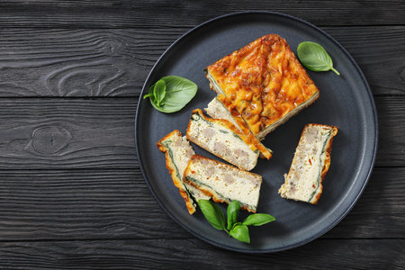 sausage egg casserole on ceramic plate, garnished with basil on black wood textured table,...