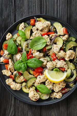 chunks of chicken breast with zucchini, bell peppers and italian seasoning sprinkled with fresh basil on a plate on black wood table, flat lay, close-up, not AI generatedの写真素材