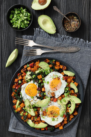 Sweet potato cubes fried and served with sunny-side-up eggs and fresh avocado slices on a plate on black wooden table with forks and napkin, vertical view, not AI-generatedの写真素材