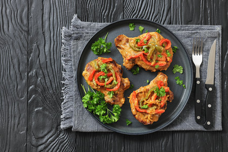 pork chops topped with bell pepper cream gravy on a plate on black wood table with fork and knife, horizontal view, flat lay, free space, real photo, not AI generatedの写真素材