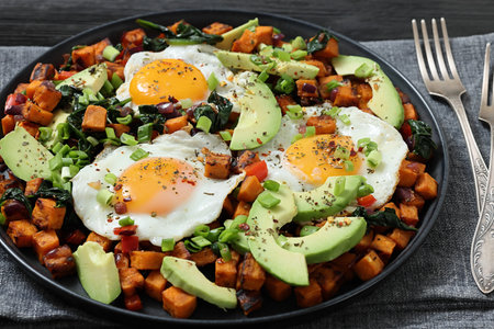 fried Sweet potato cubes served with sunny-side-up eggs and fresh avocado slices on a plate on black wooden table, close-up, not AI-generatedの写真素材