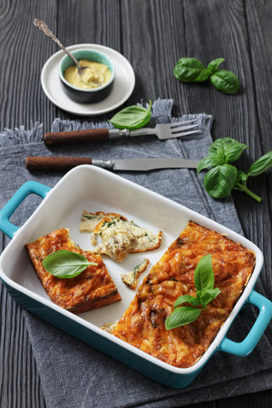 Sausage and cheesy egg breakfast casserole in a blue dish on wooden table with basil and mustard, vertical view, authentic photoの写真素材