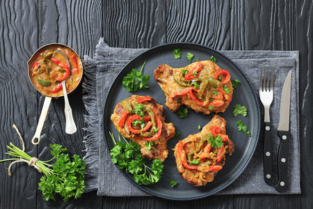 pork chops topped with bell pepper cream gravy on a plate on black wood table with parsley, fork and knife, horizontal view, flat lay, free space, real photo, not AI generatedの写真素材