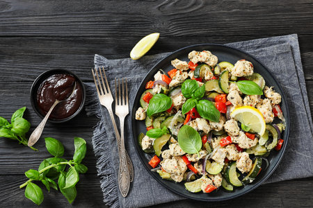 chunks of chicken breast with zucchini, bell peppers and italian seasoning sprinkled with fresh basil on a plate on black wood table with forks, flat lay, not AI generatedの写真素材
