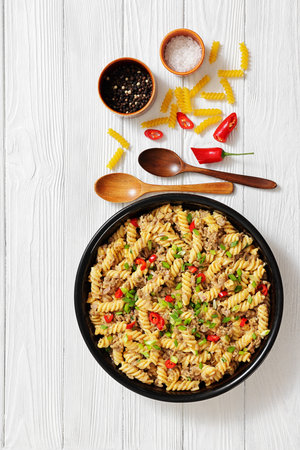 creamy ground beef pasta, cheeseburger fusilli pasta sprinkled with green onion and chili pepper in a bowl on white wood table with spoons and ingredients, vertical view, not AI generatedの写真素材