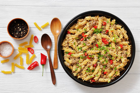 creamy ground beef pasta, cheeseburger fusilli pasta sprinkled with green onion and chili pepper in a bowl on white wood table with spoons and ingredients, flat lay, not AI generatedの写真素材