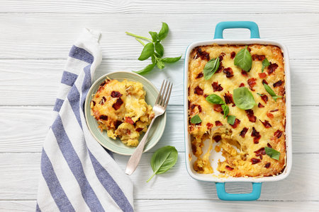 twice baked potato casserole with fried bacon and cheddar cheese in a baking dish and a portion on a plate on white wood table, horizontal view, flat lay, not AI generatedの写真素材