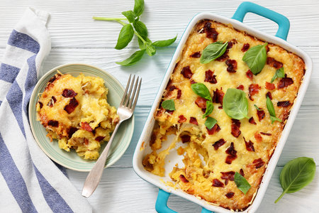 twice baked potato casserole with fried bacon and cheddar cheese in a baking dish and a portion on a plate on white wood table, horizontal view, flat lay, close-up, not AI generatedの写真素材