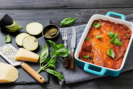 Eggplant Parmigiana, slices of eggplant layered with marinara sauce, parmesan and mozzarella cheese in a baking dish on black wood table with ingredients and cutlery,  not AI generatedの写真素材
