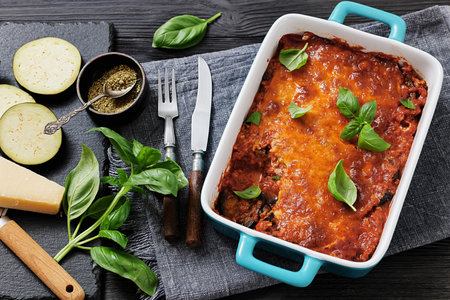 Eggplant Parmigiana, slices of eggplant layered with marinara sauce, parmesan and mozzarella cheese in a baking dish on black wood table with ingredients and cutlery,  flat lay, not AI generatedの写真素材