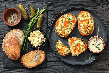 deviled egg salad and red caviar on toast served on a plate with mayo mustard sauce on black wood table with ingredients and dill pickles, horizontal view, flat lay, close-up, not AI generatedの写真素材